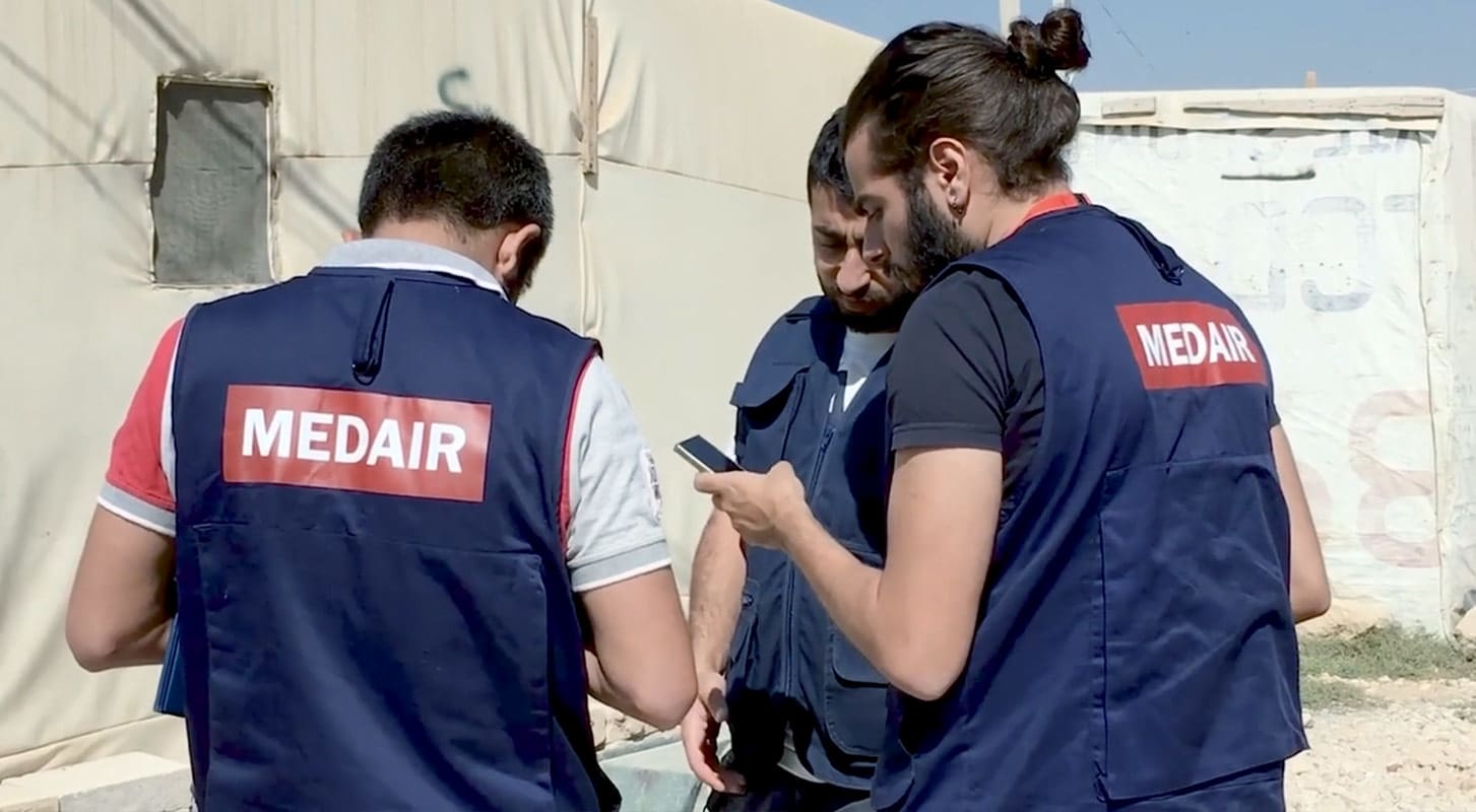 Three men wearing Medair vests converse next to a tent, each looking at their phones.